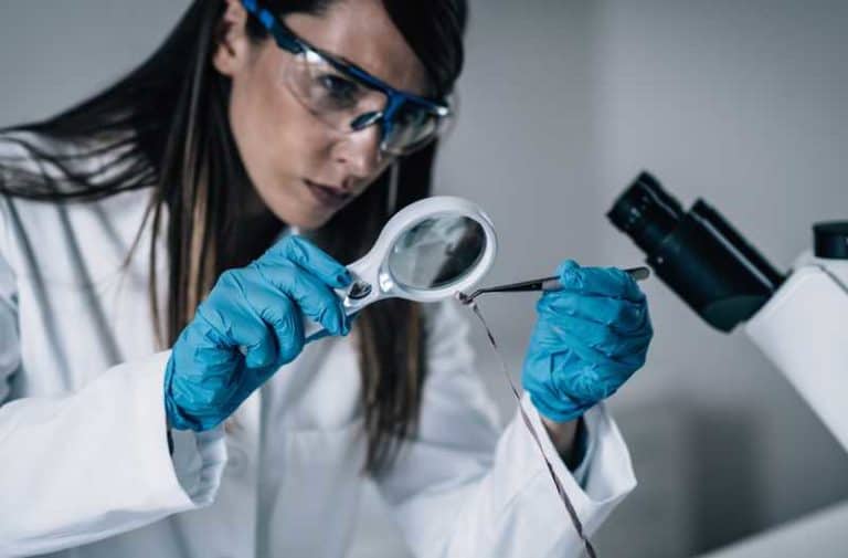 Een vrouw in laboratoriumjas en veiligheidsbril houdt een klein glazen plaatje vast met een pincet