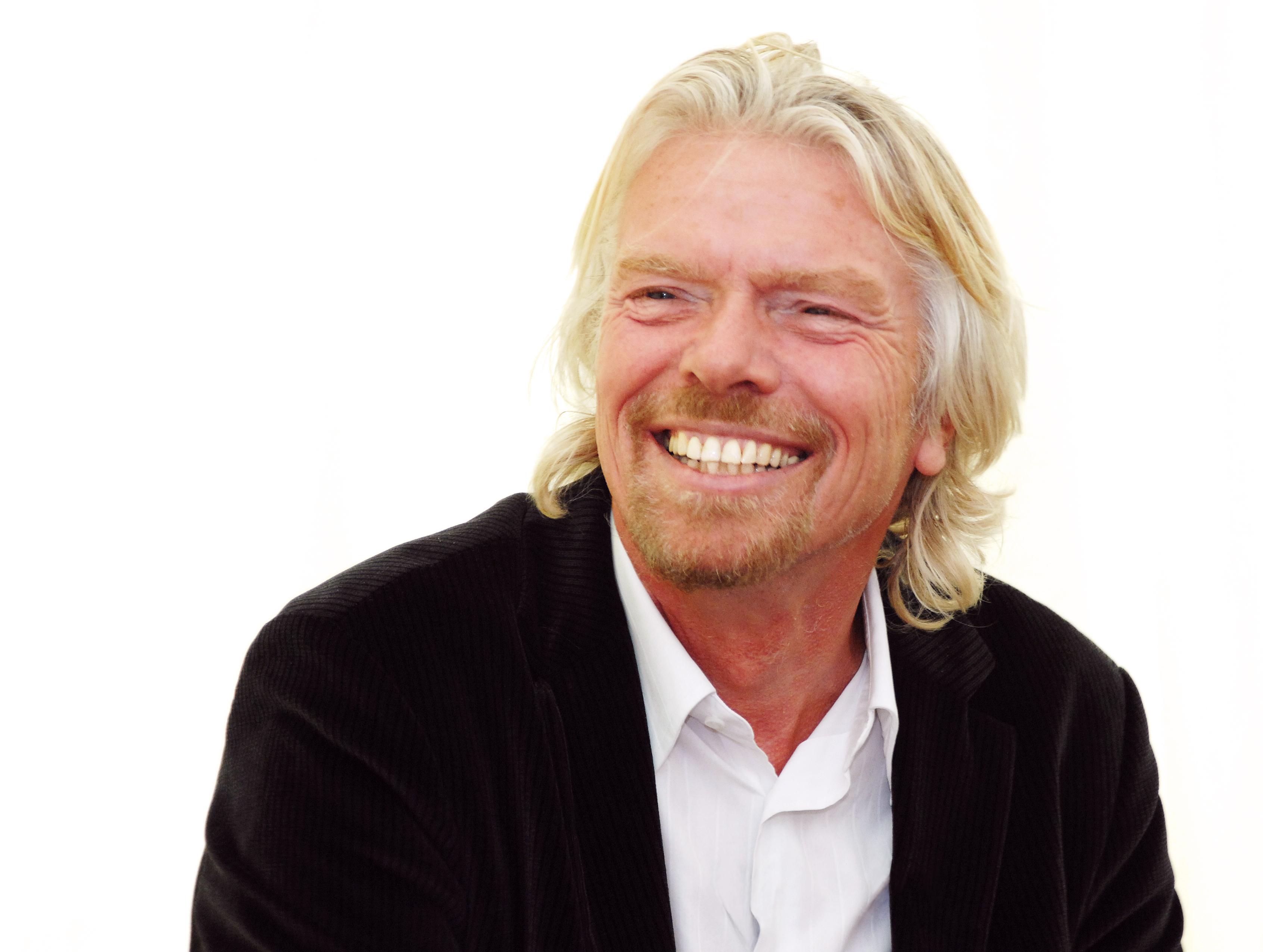 Een lachende man met blond haar in een zwart pak en wit overhemd (Richard Branson)