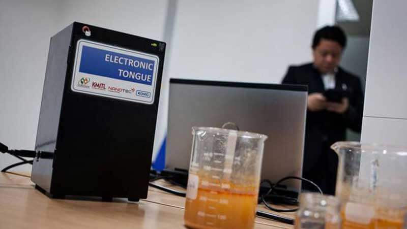 Twee glazen reageerbuizen met oranje vloeistof staan op een tafel naast een zwart apparaat met het woord ‘electronic tongue’ met op de achtergrond een man