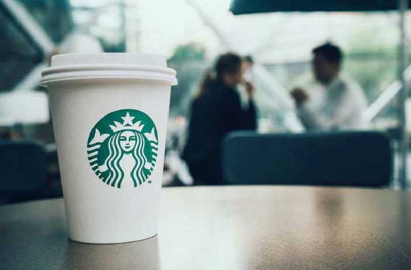 Een Starbucks koffiebeker staat op een houten tafel met op de achtergrond twee mensen die met elkaar praten