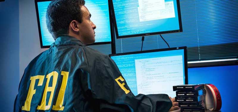 Persoon met een FBI-jas zit en kijkt naar drie computerschermen