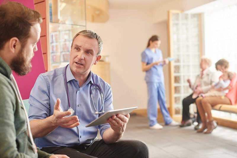 Een dokter houdt een tablet vast en spreekt tegen een man in een ziekenhuisgang