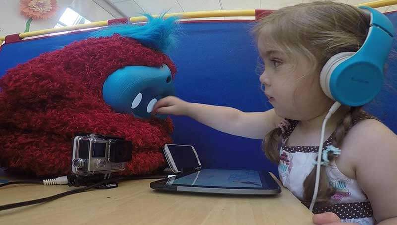 Een meisje met blauwe hoofdtelefoon speelt met robotspeelgoed met voor haar een smartphone en een tablet