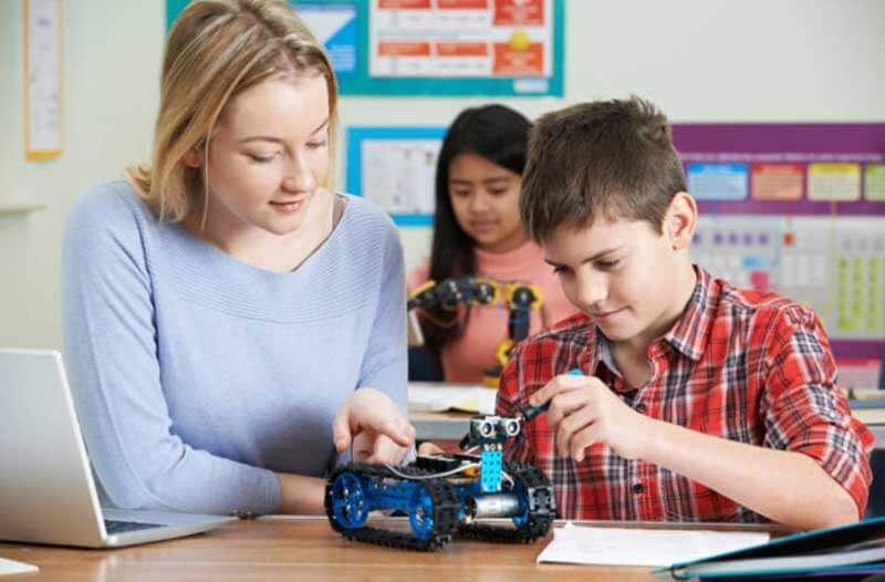 Two primary school pupils sitting in a classroom analysing small robots with the help of a teacher