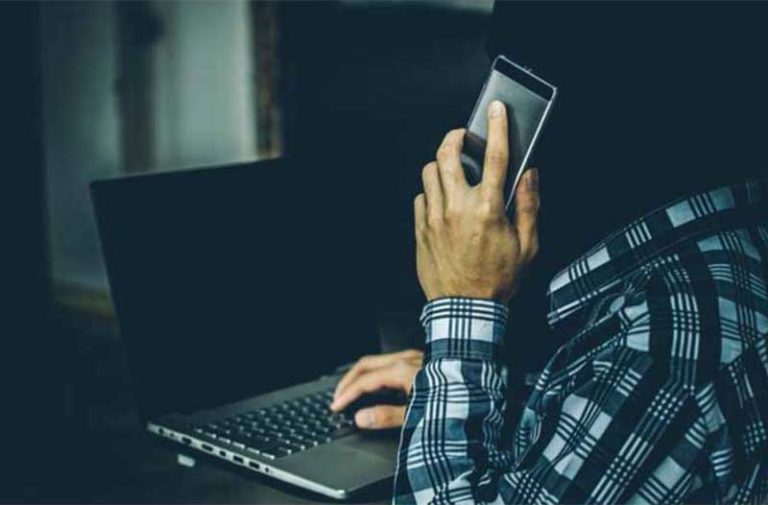 Man met geruit shirt typt op een laptop en houdt een smartphone in zijn linkerhand