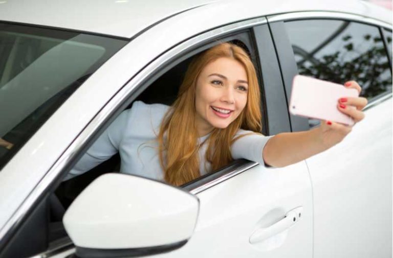 Een vrouw met lang oranje haar zit in een witte auto en neemt een selfie