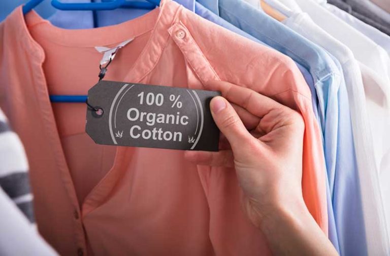 Een hand houdt een label vast met ‘100% organic cotton’ dat aan een roze shirt bevestigd is|||A hand holding a “100% organic cotton” certification label on a peach-coloured shirt