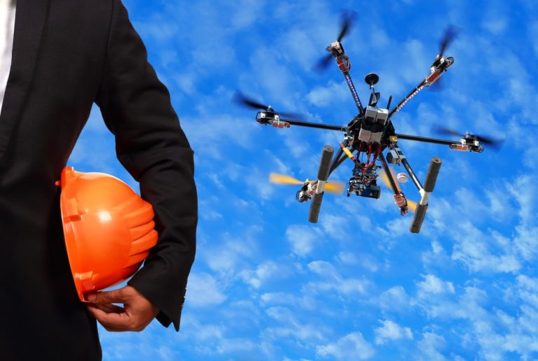 Een man in een zwart pak en een oranje helm onder zijn arm staat naast een drone die in een blauwe lucht vliegt