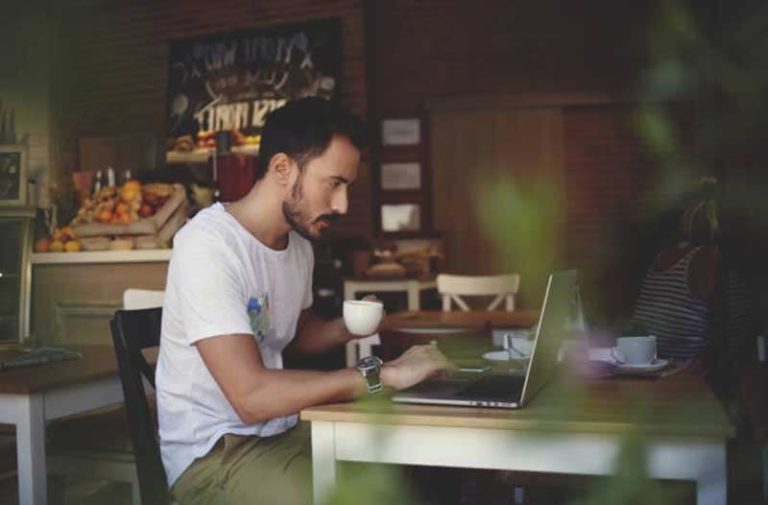 Een man met wit t-shirt zit in een cafeetje koffie te drinken terwijl hij op zijn laptop werkt