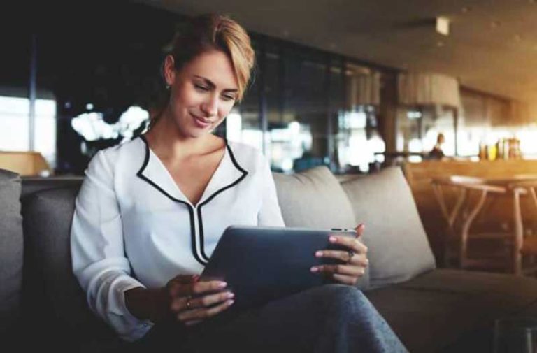 Een blonde vrouw met een wit shirt zit op een bank en kijkt op haar tablet