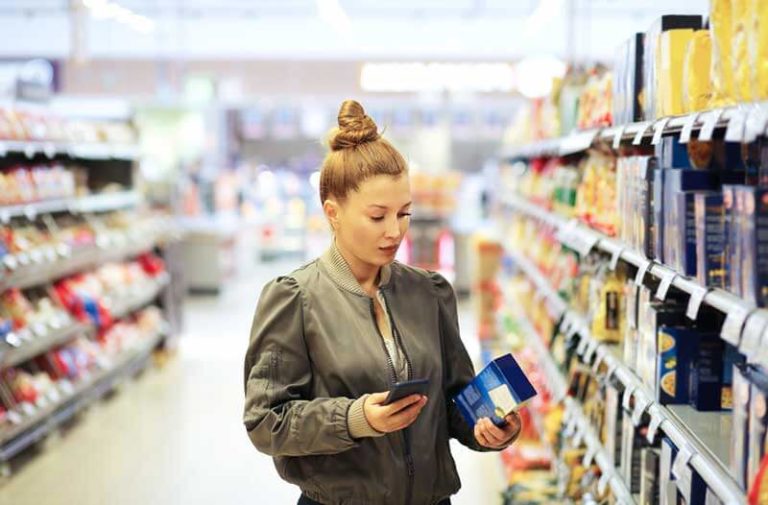 Een vrouw staat in een winkel met een artikel in het ene hand en een smartphone in de andere