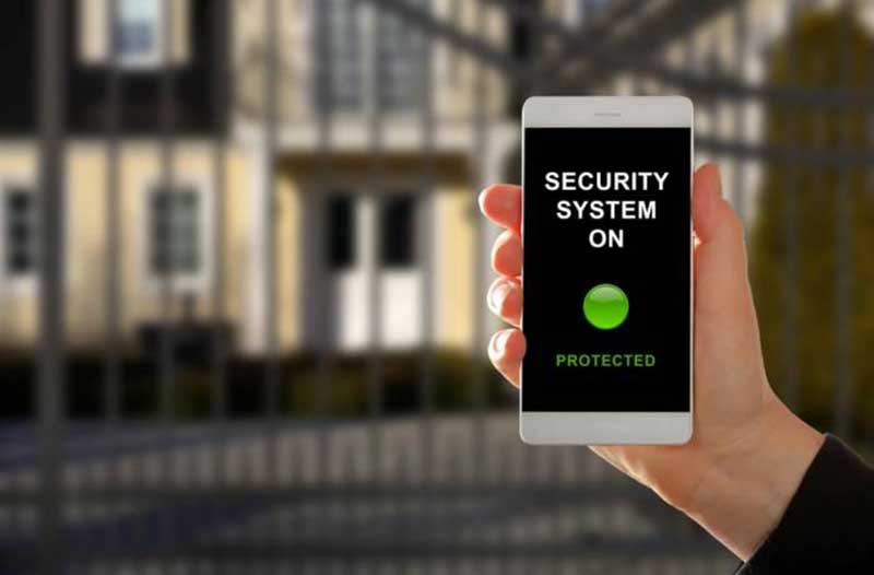 Hand holding a smartphone displaying information that the security system is on, with a house gate in the background
