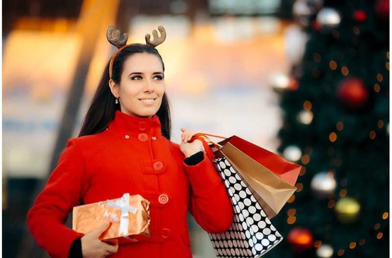 Een vrouw met lang donker haar, rode jas, rendiergewei op haar hoofd en kerstinkopen in tassen in haar handen loopt langs een kerstboom