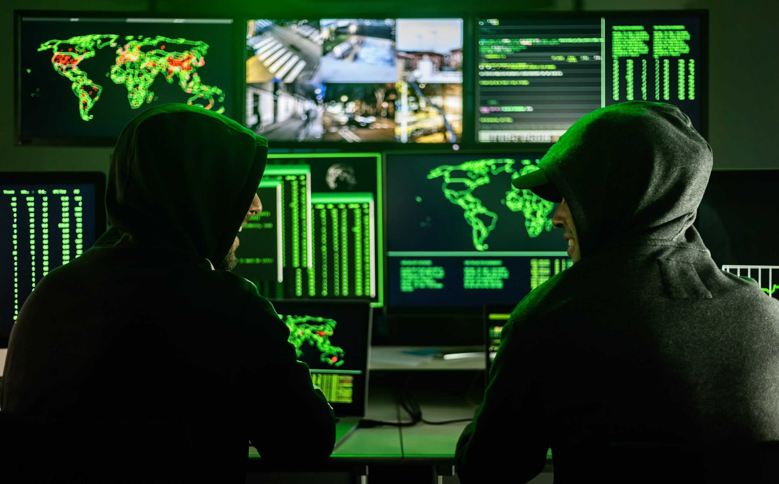 Two people with black hoodies are sitting in a dark room in front of a huge screen that shows maps of the world and images of street corners and buildings.