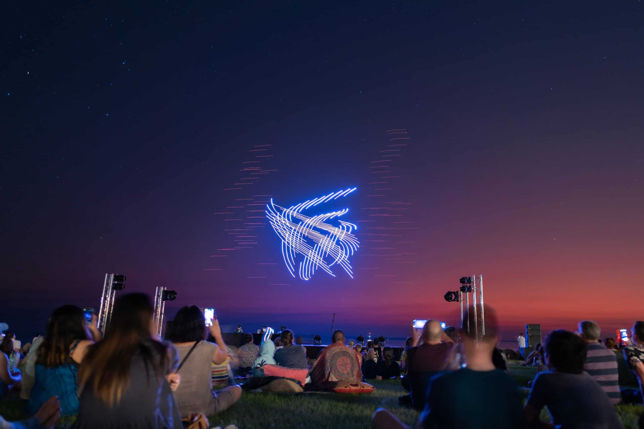 A dark sky illuminated by a drone show, with people in the audience capturing videos and photos on their mobile phones.