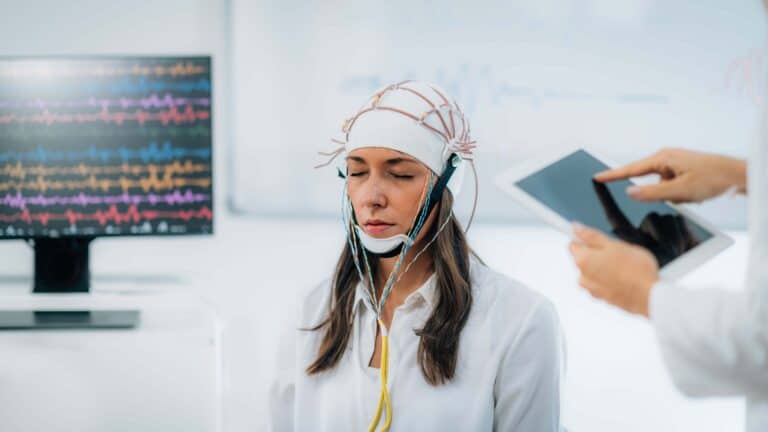 Een vrouw krijgt een EEG waarvoor elektroden op haar hoofd zijn aangebracht. Daarnaast staat een zorgverlener die een tablet vasthoudt.
