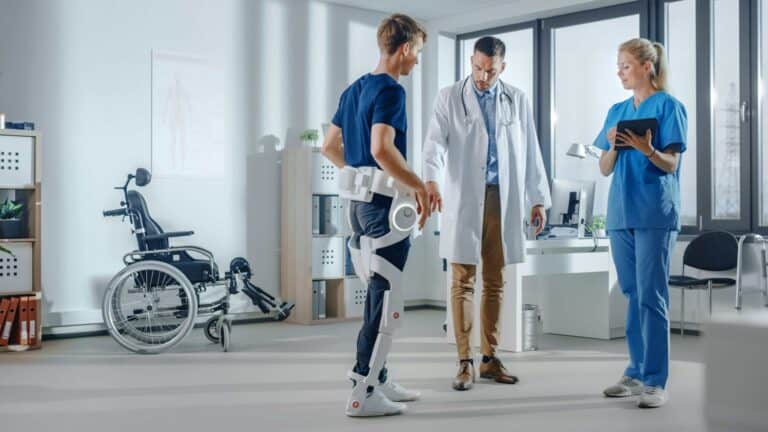 Een patiënt in een been-exoskelet wordt bijgestaan door medisch personeel in een kliniek, met een rolstoel op de achtergrond.