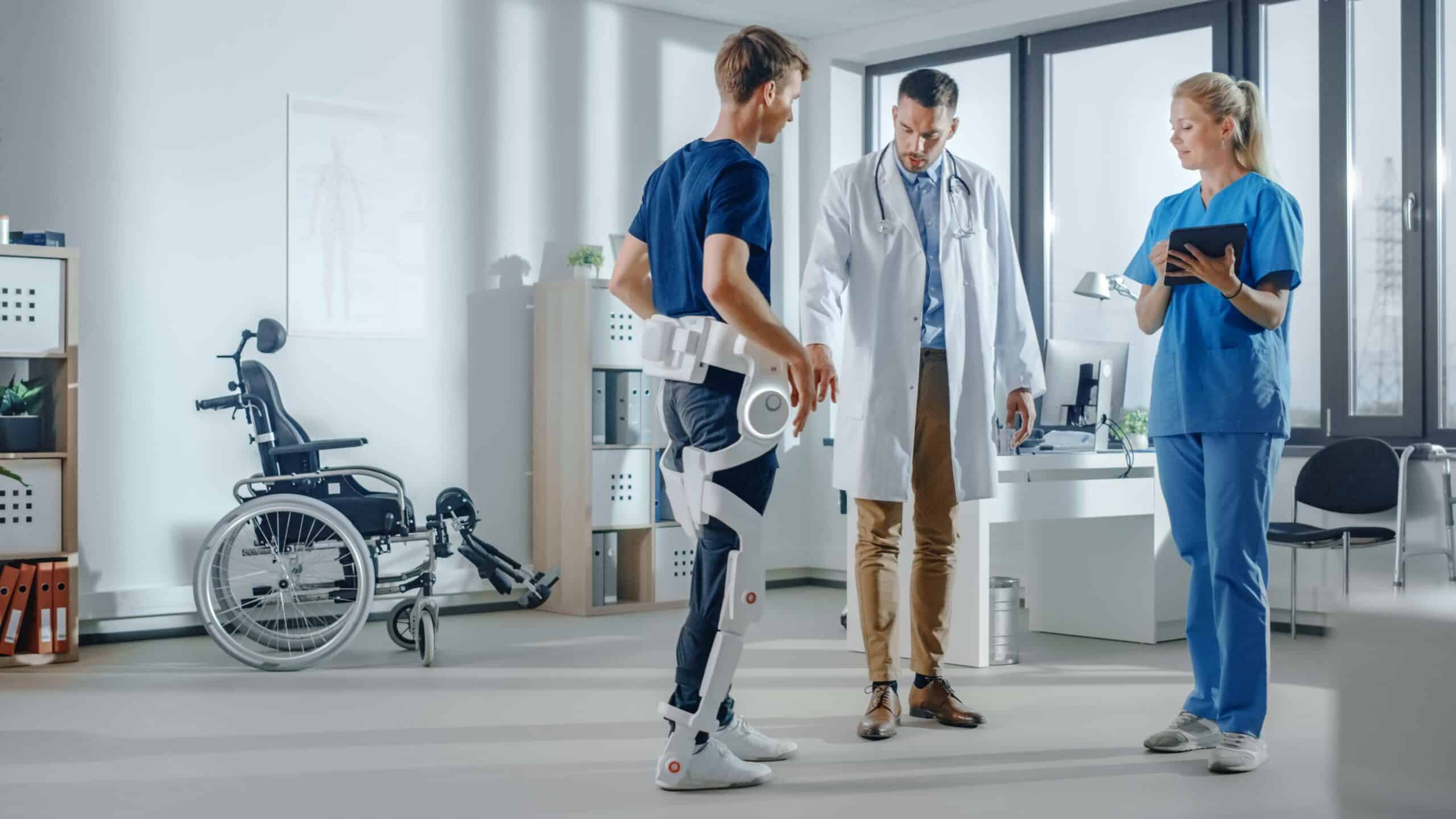 A patient in a leg exoskeleton is assisted by medical staff in a clinic, with a wheelchair in the background.