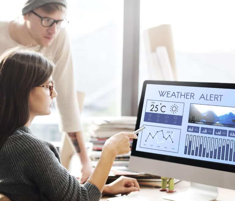 Een vrouw wijst naar een computerscherm met een weeralarm, terwijl een man achter haar meekijkt.