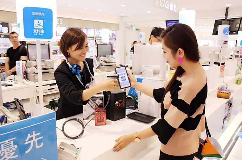 A girl holding her smartphone and using Alipay in a store while a female cashier is scanning her smartphone and smiling