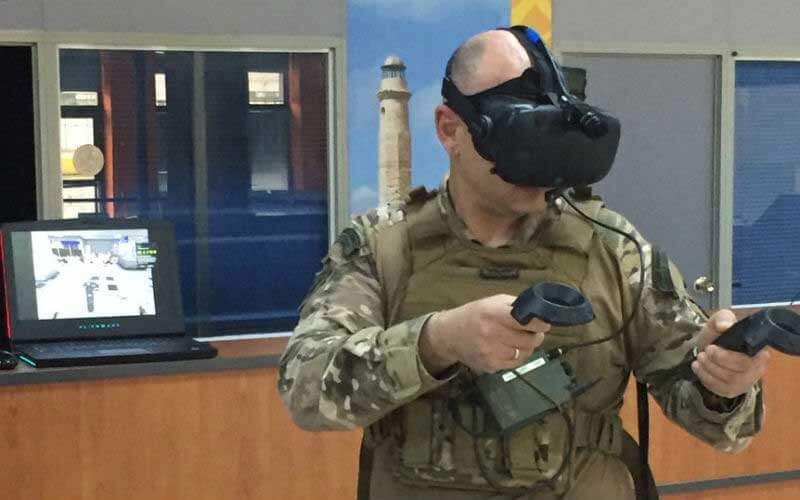  A soldier wearing a VR headset and holding VR controllers, with a laptop in the background 