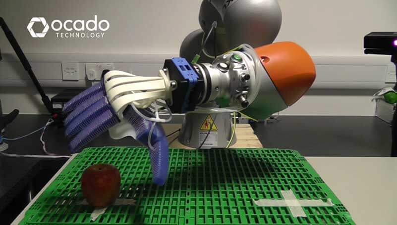 A robotic arm with flexible fingers grabbing an apple from a green surface 