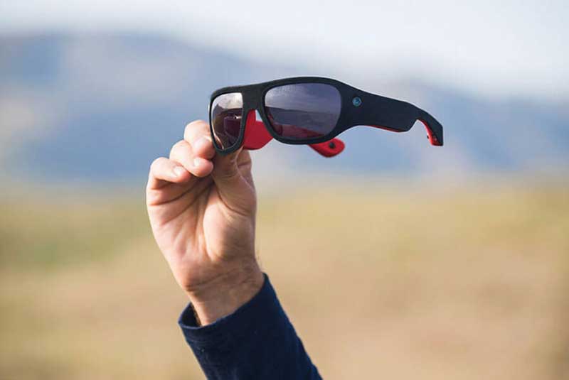 A hand holding sunglasses with a field blurred in the background
