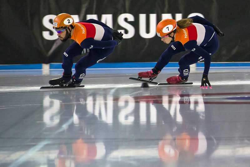  Two speed skaters side-by-side on ice with Samsung’s logo in the background 