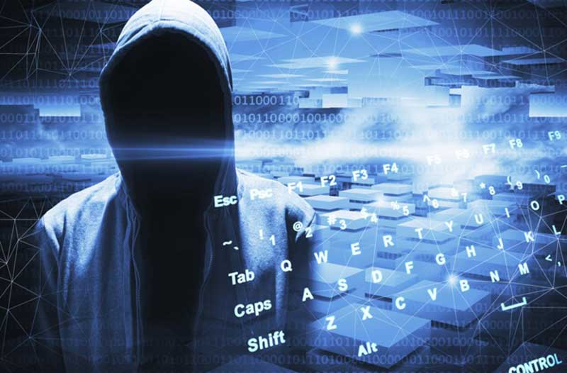 A person in a hoodie with their face covered, with a digital computer keyboard next to them and lines of code in the background. 