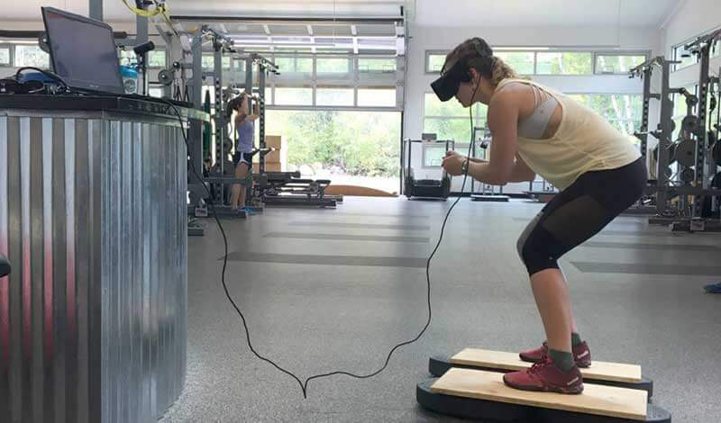 A woman in the gym wearing a VR headset attached to a laptop in front of her