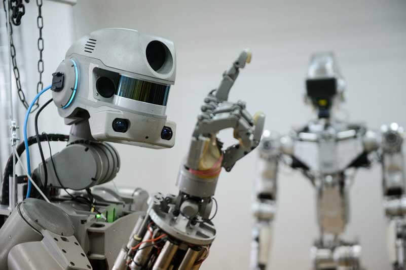 A robot holding his index finger up, while another robot is standing in the background