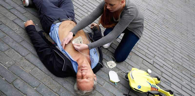 A man lying unconscious on the ground while a woman is attaching the defibrillator to his chest