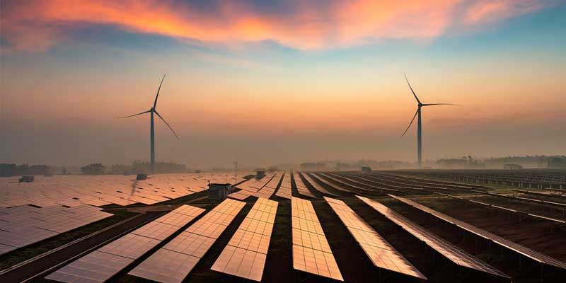 A solar farm with two wind turbines in the background at sunset