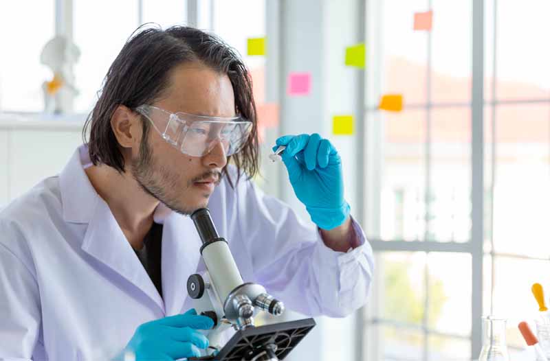 Man in white laboratory suit wearing protective spectacles looking at something through a microscope