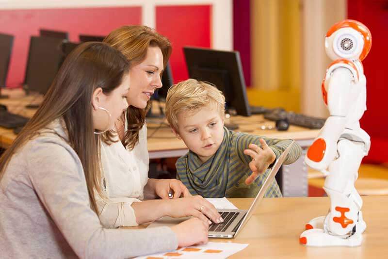 Robot standing in front of two female teachers and a boy working together on a laptop