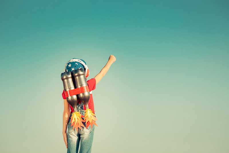 Child with red t-shirt, jeans, cap, and headphones with toy rockets strapped to his back stretching out his arm and making a fist