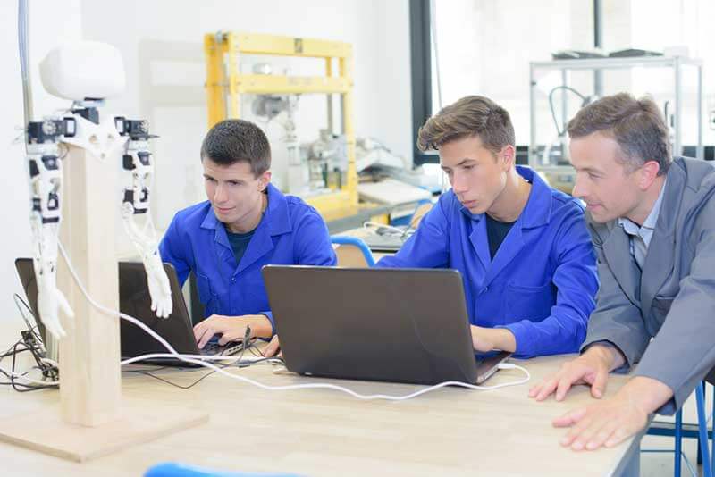 A supervisor observes two student trainees working on computers in a robotics lab