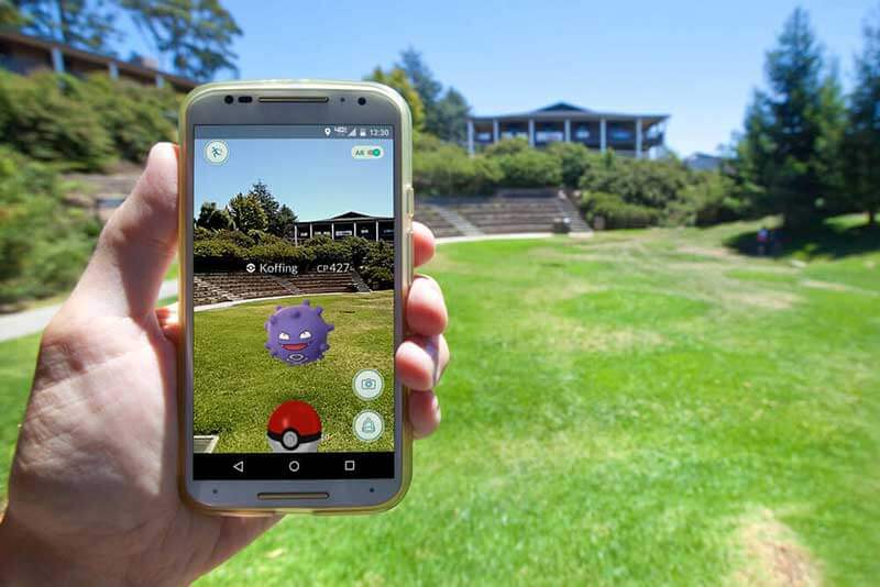 Hand holding a smartphone with Pokemon Go outdoors with greenery and a building in the background