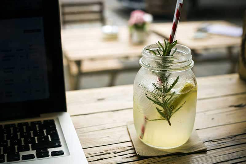 A glass with a cold drink and a straw on a wooden table with a laptop next to it